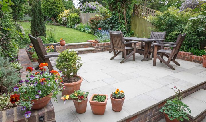 A raised patio garden with wood furniture set and brick retaining walls, surrounded with landscaped yard